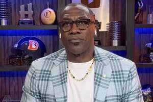 Person wearing glasses and a plaid blazer with a white shirt, in a room with sports memorabilia, possibly during an interview or broadcast