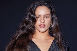 Person posing in an elegant, black textured outfit against a plain backdrop, with long wavy hair framing their face